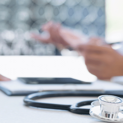 Closeup of a doctor's hands holding a pen over a table with stethoscope and paper on it, representing benefits of interventional radiology, treatment for varicose veins