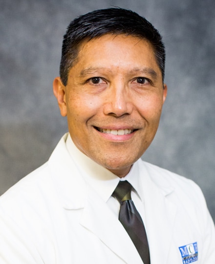 Alan fontanares. Professional headshot of a medical provider wearing a white coat and tie against a gray studio background.