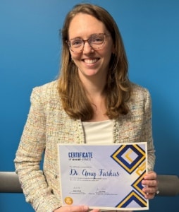 Dr. Amy farkas smiling and holding an award certificate in front of a blue wall.
