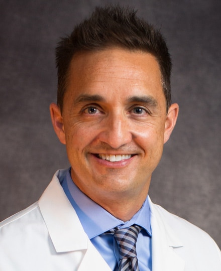 Chris dobzyniak, md. Portrait of a male health professional in a white coat and patterned tie against a professional, gray backdrop.