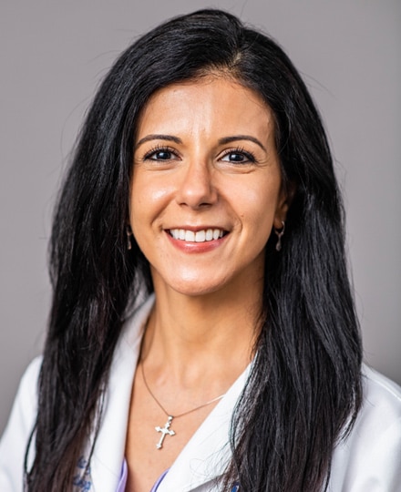 Dina elgohary, md. Professional headshot of a female radiologist with long dark hair wearing a white medical coat and a cross necklace, smiling against a neutral gray background.