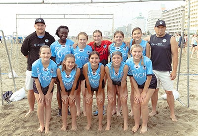 Girls youth beach soccer team wearing light blue mcr jerseys posing with their coaches at a tournament.