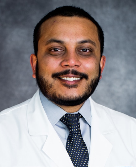 Meet dr. Krishna chaudhuri, md, pr. Professional headshot of a male radiologist wearing a white medical coat and patterned tie, smiling in front of a neutral studio background.