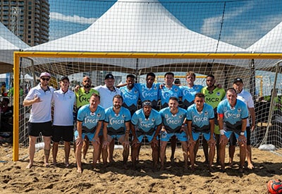 Men’s beach soccer team wearing blue mcr jerseys posing together in front of a goal at a beach tournament.