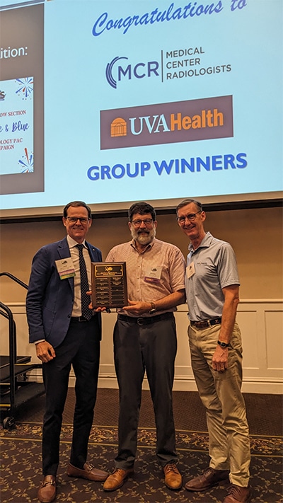 Three representatives from medical center radiologists and uva health accept a group award at a radpac event, holding a plaque in front of a presentation screen announcing the winners.