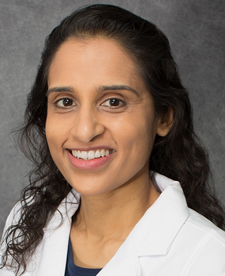 Professional headshot of a female radiologist with long dark curly hair wearing a white medical coat, smiling in front of a neutral gray background.