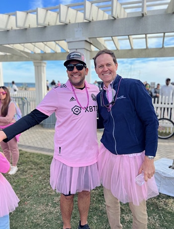 Two men wearing pink tutus and pink-themed clothing smiling together at a breast cancer awareness walk event.