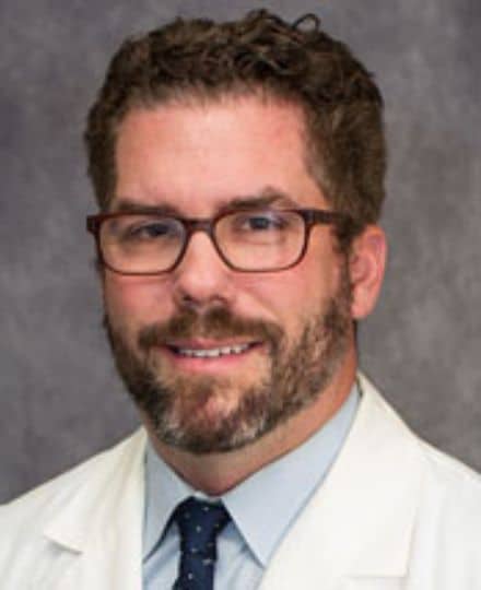 Travis dozier, pa. Male medical professional wearing glasses, a dress shirt, tie, and white lab coat, posing for a formal headshot.